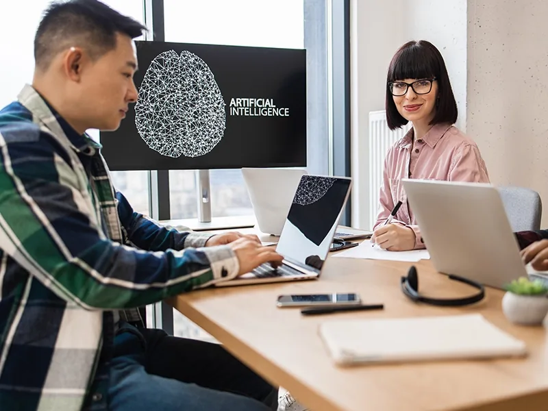 A team working at laptops in an office, an AI model on the screen behind them